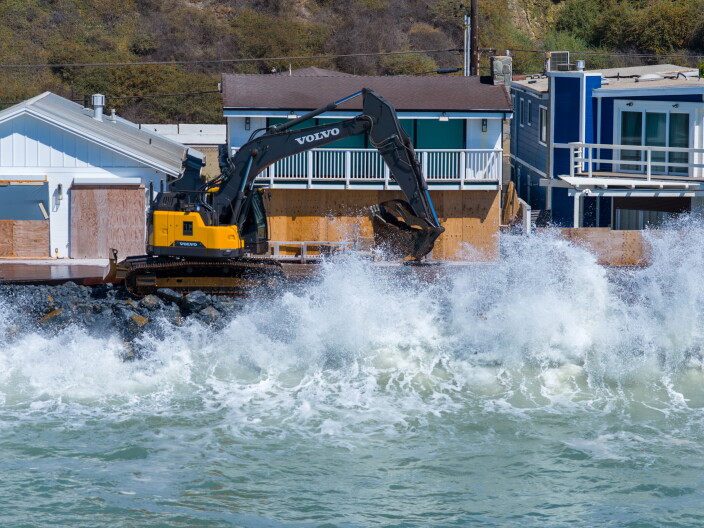 An excavator places rocks along a seawall as large waves crash against beachfront homes.